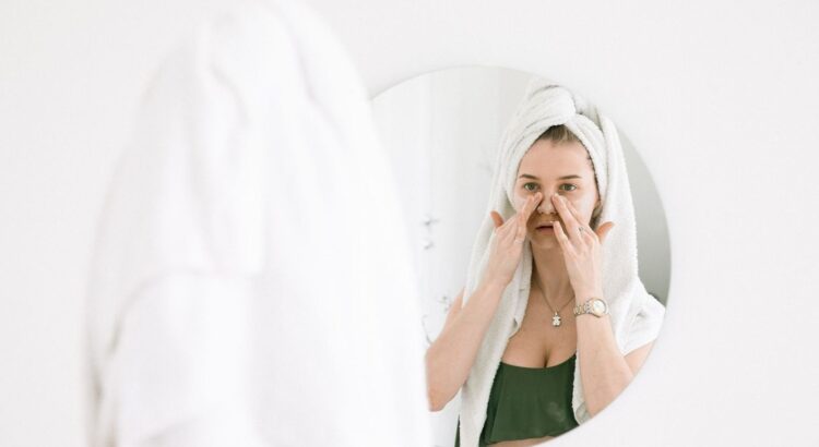 Woman with a towel-wrapped head applying skincare in a bathroom mirror